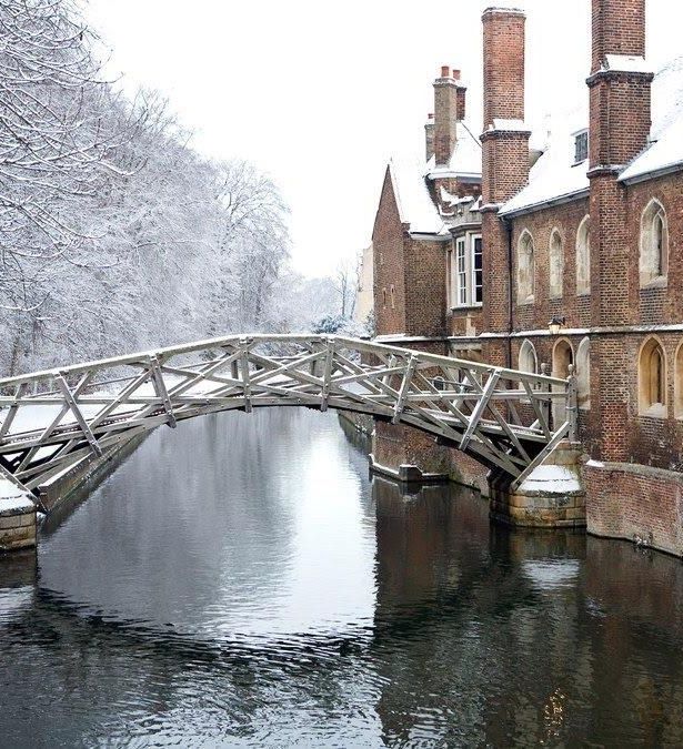 Wooden Mathematical Bridge covered in snow over the River Cam at Cambridge, with historical brick buildings and snowy trees.