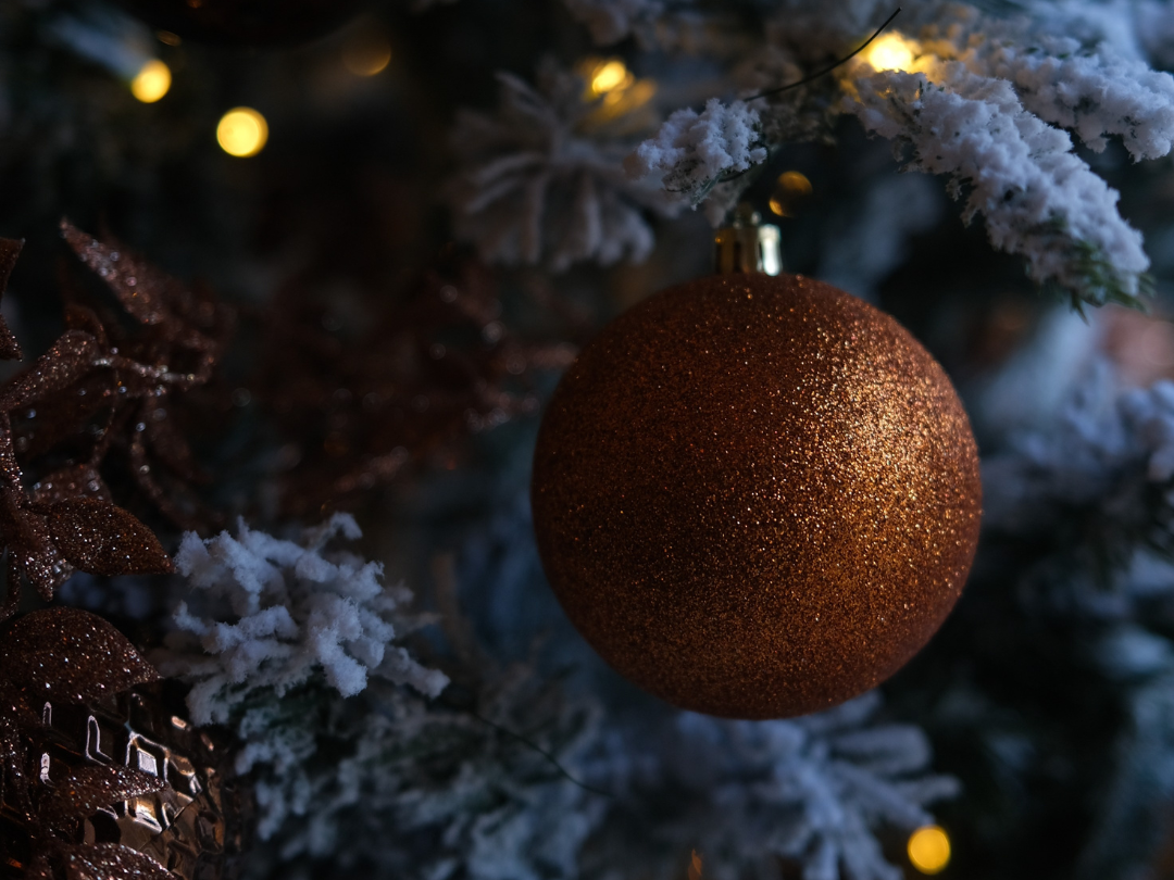 Close-up of a glittery bronze Christmas ornament hanging on a snow-frosted tree with warm lights.