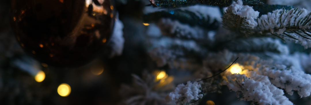 Close-up of a glittery bronze Christmas ornament hanging on a snow-frosted tree with warm lights.