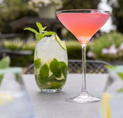 A pink cocktail in a martini glass next to a mojito garnished with mint leaves on an outdoor table.