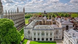 Aerial view of historic Cambridge University buildings with Gothic spires, lush green lawns, and trees under a partly cloudy sky.