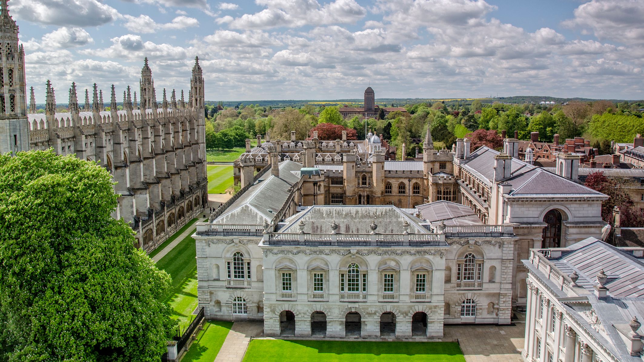 Aerial view of historic Cambridge University buildings with Gothic spires, lush green lawns, and trees under a partly cloudy sky.