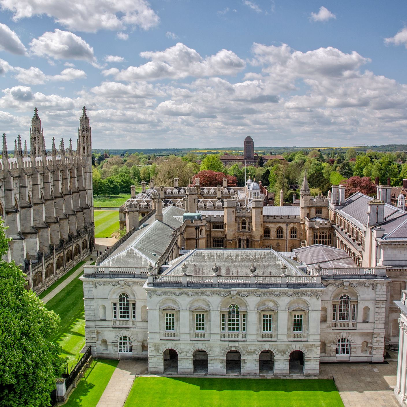 Aerial view of historic Cambridge University buildings with Gothic spires, lush green lawns, and trees under a partly cloudy sky.