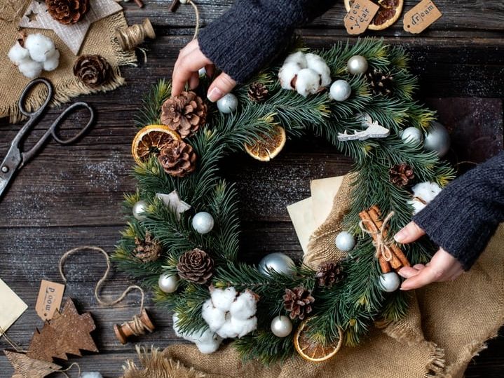 Person arranging a festive Christmas wreath decorated with pinecones, cotton, dried orange slices, and ornaments on a rustic wooden table.