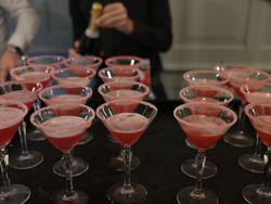 Several pink cocktails in sugar-rimmed martini glasses arranged on a tray, with people preparing drinks in the background.