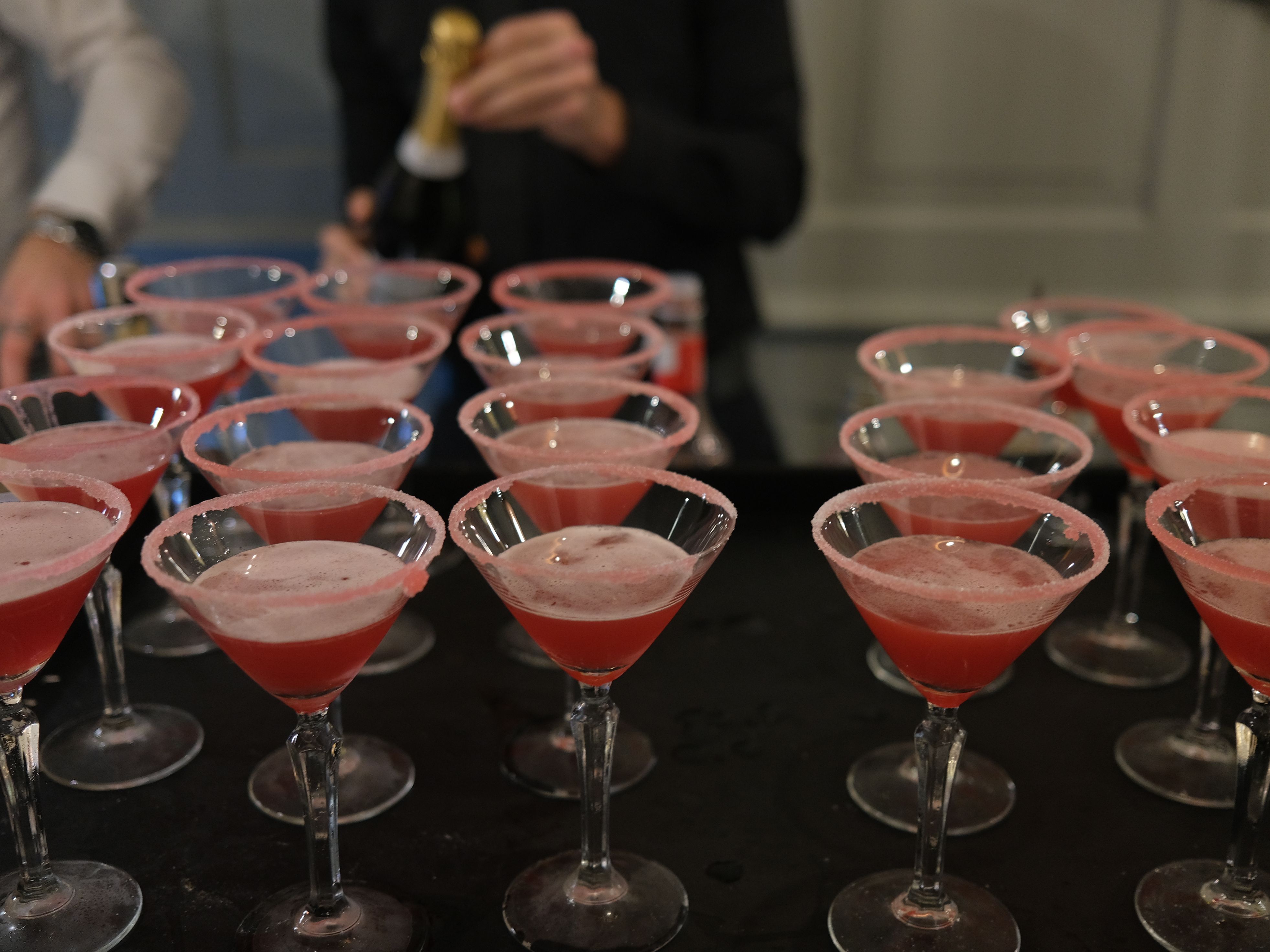 Several pink cocktails in sugar-rimmed martini glasses arranged on a tray, with people preparing drinks in the background.