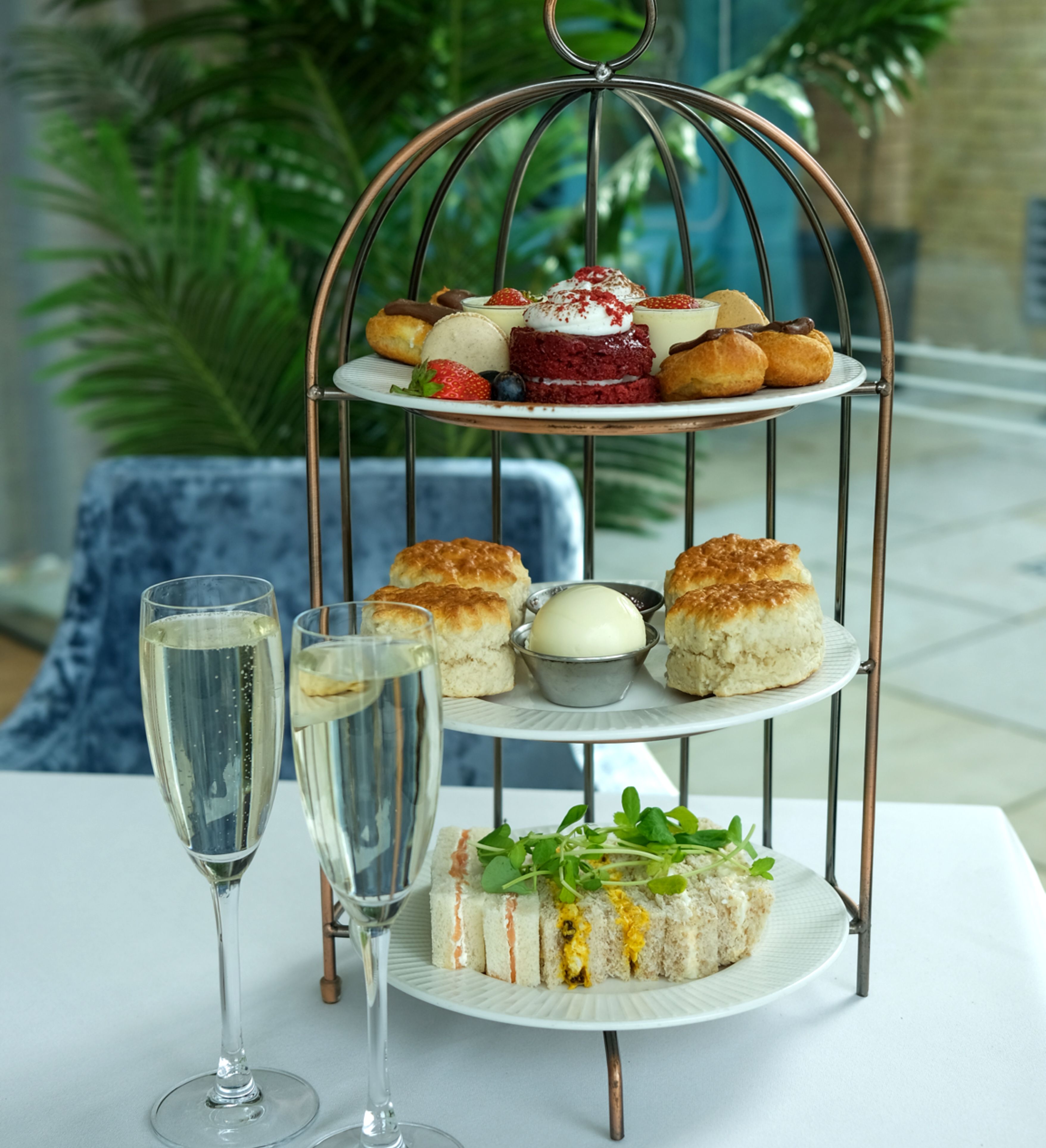 Three-tiered stand with pastries, scones, and sandwiches on a table, accompanied by two glasses of champagne.