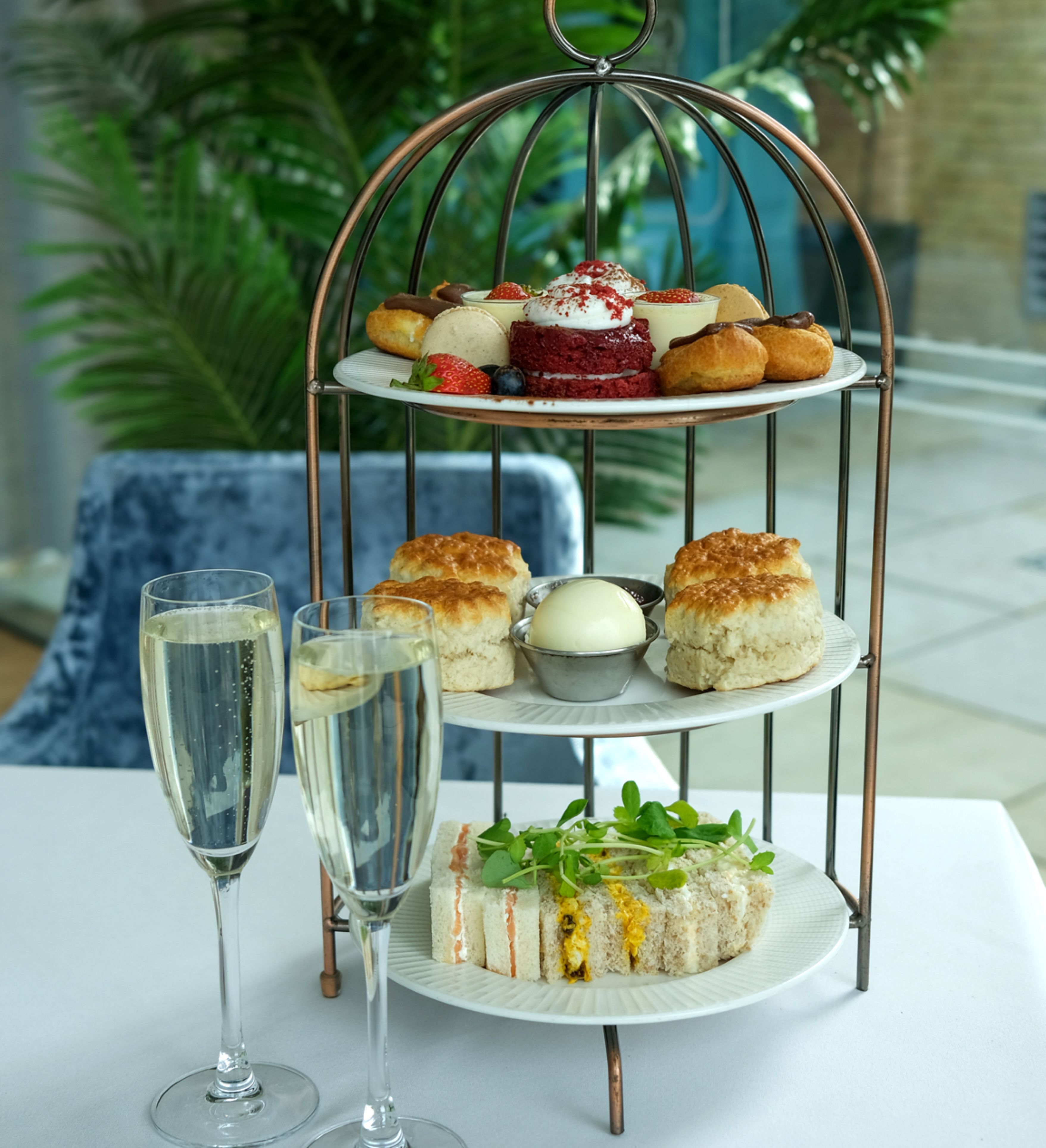 Three-tiered stand with pastries, scones, and sandwiches on a table, accompanied by two glasses of champagne.