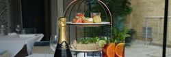 Afternoon tea set with sandwiches, scones, pastries, a bottle of sparkling wine, and two glasses of fruit cocktails on a restaurant table.