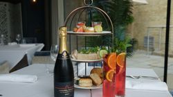 Afternoon tea set with sandwiches, scones, pastries, a bottle of sparkling wine, and two glasses of fruit cocktails on a restaurant table.