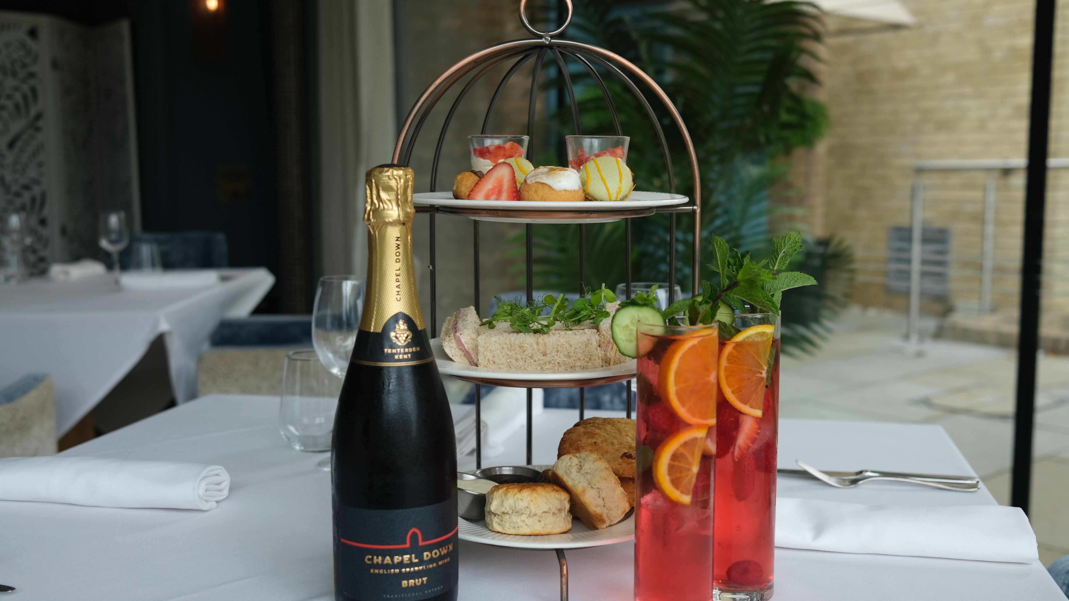 Afternoon tea set with sandwiches, scones, pastries, a bottle of sparkling wine, and two glasses of fruit cocktails on a restaurant table.