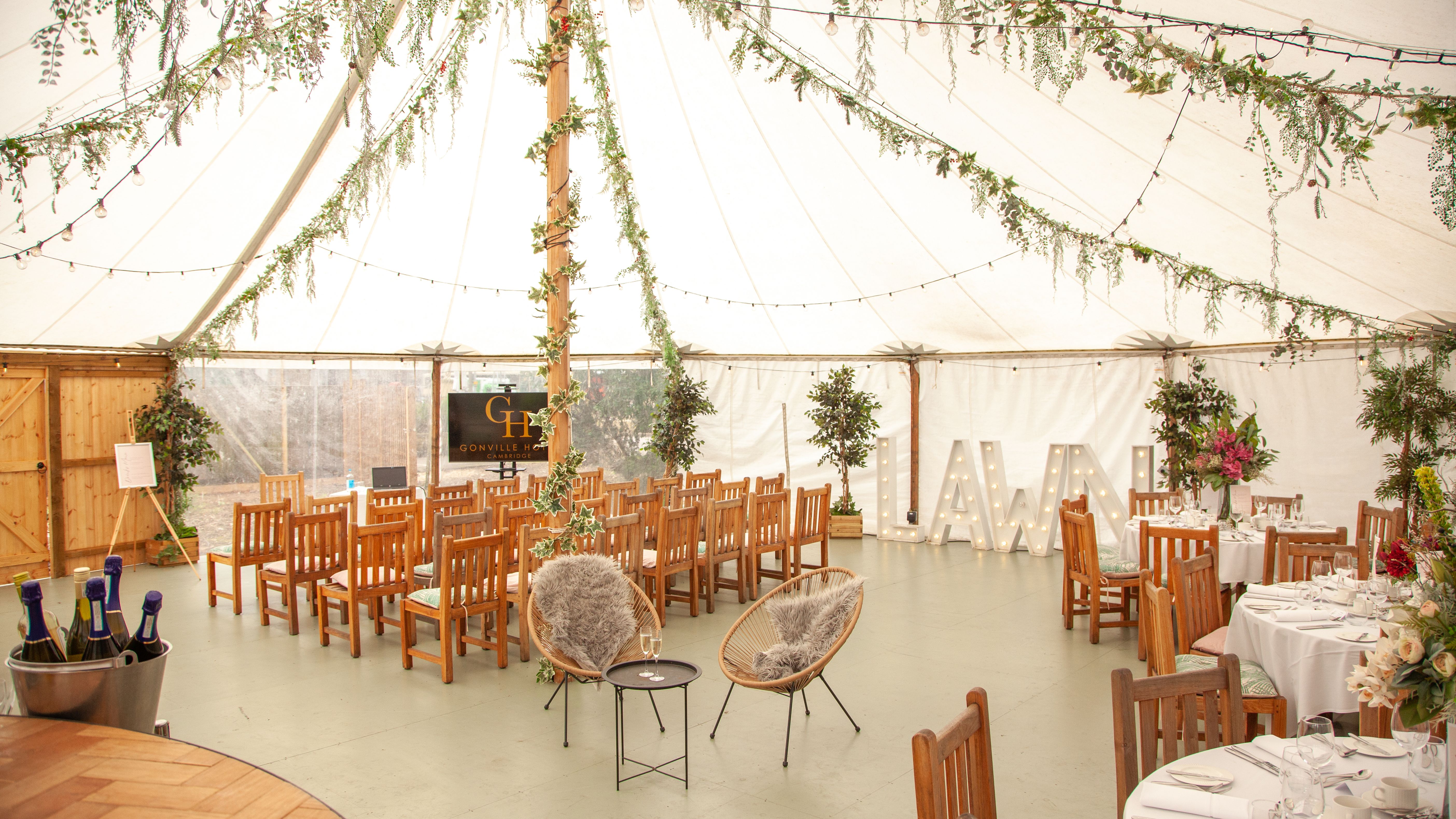 Decorated teepee event tent interior with wooden chairs, round dining tables, string lights, and greenery.