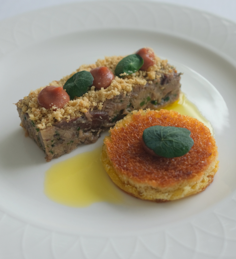 A gourmet plated dish with a rectangular meat terrine topped with crumbs and herbs, next to a round golden crusted savory item, garnished with green leaves.