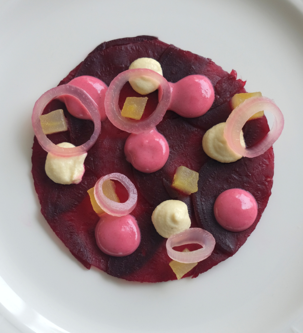 Artistic beetroot carpaccio garnished with dollops of mousse, pickled onion rings, and small cubes on a white plate