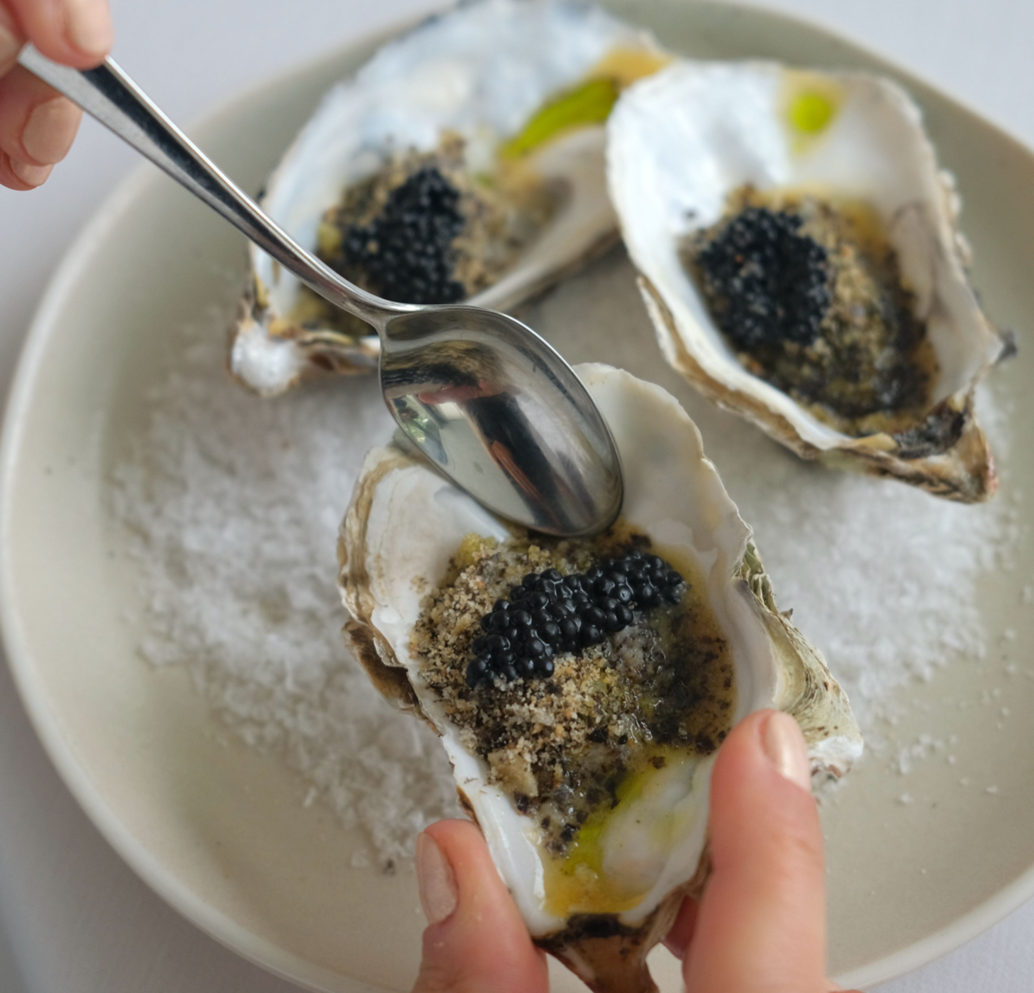 Close-up of a hand holding an oyster shell with black caviar, breadcrumbs, and a drizzle of oil, on a plate with more oysters and sea salt.