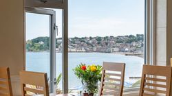 Dining table set by a large window with a scenic view of a coastal town and water, with flowers in a vase at the center of the table.