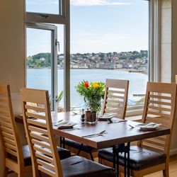 Dining table set by a large window with a scenic view of a coastal town and water, with flowers in a vase at the center of the table.