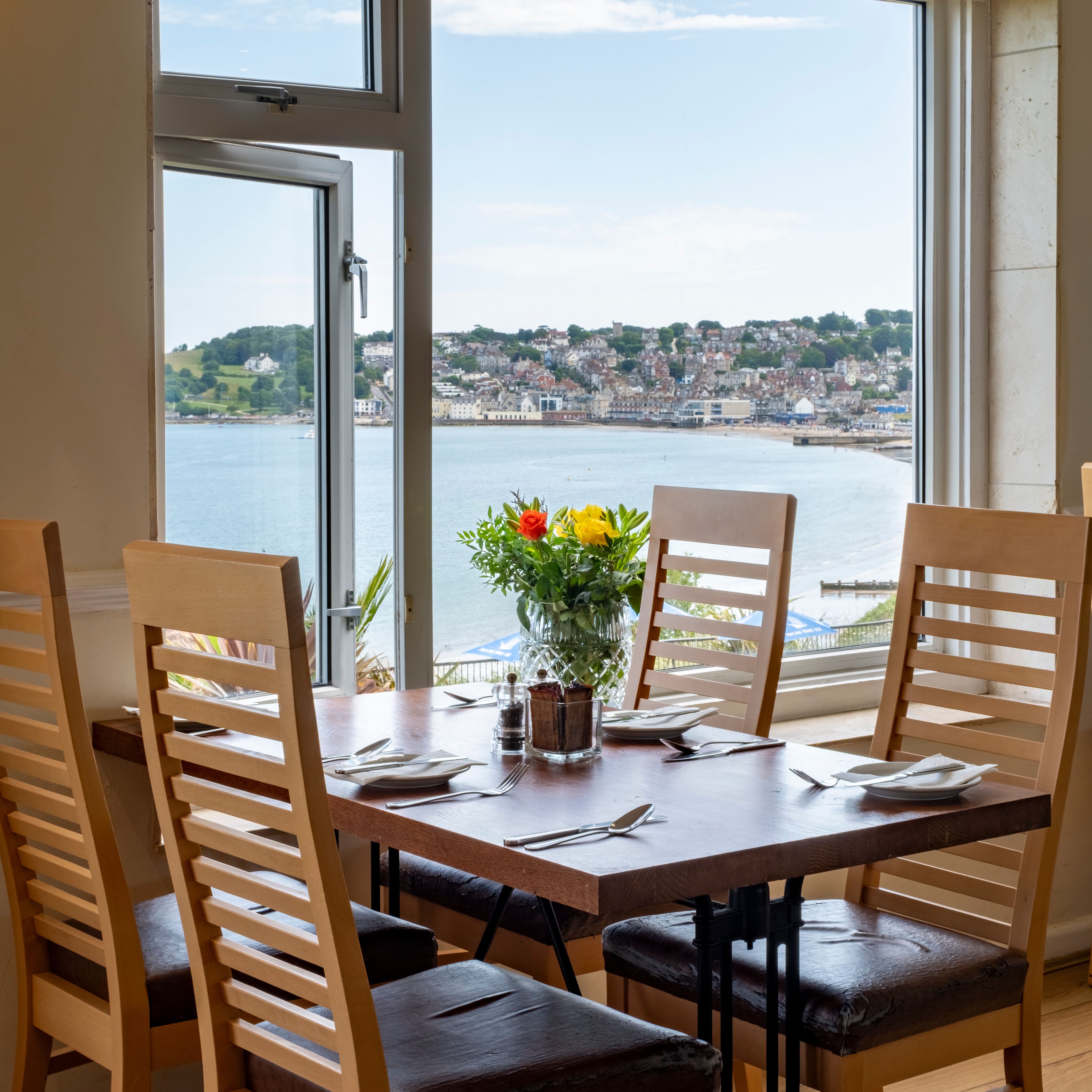 Dining table set by a large window with a scenic view of a coastal town and water, with flowers in a vase at the center of the table.