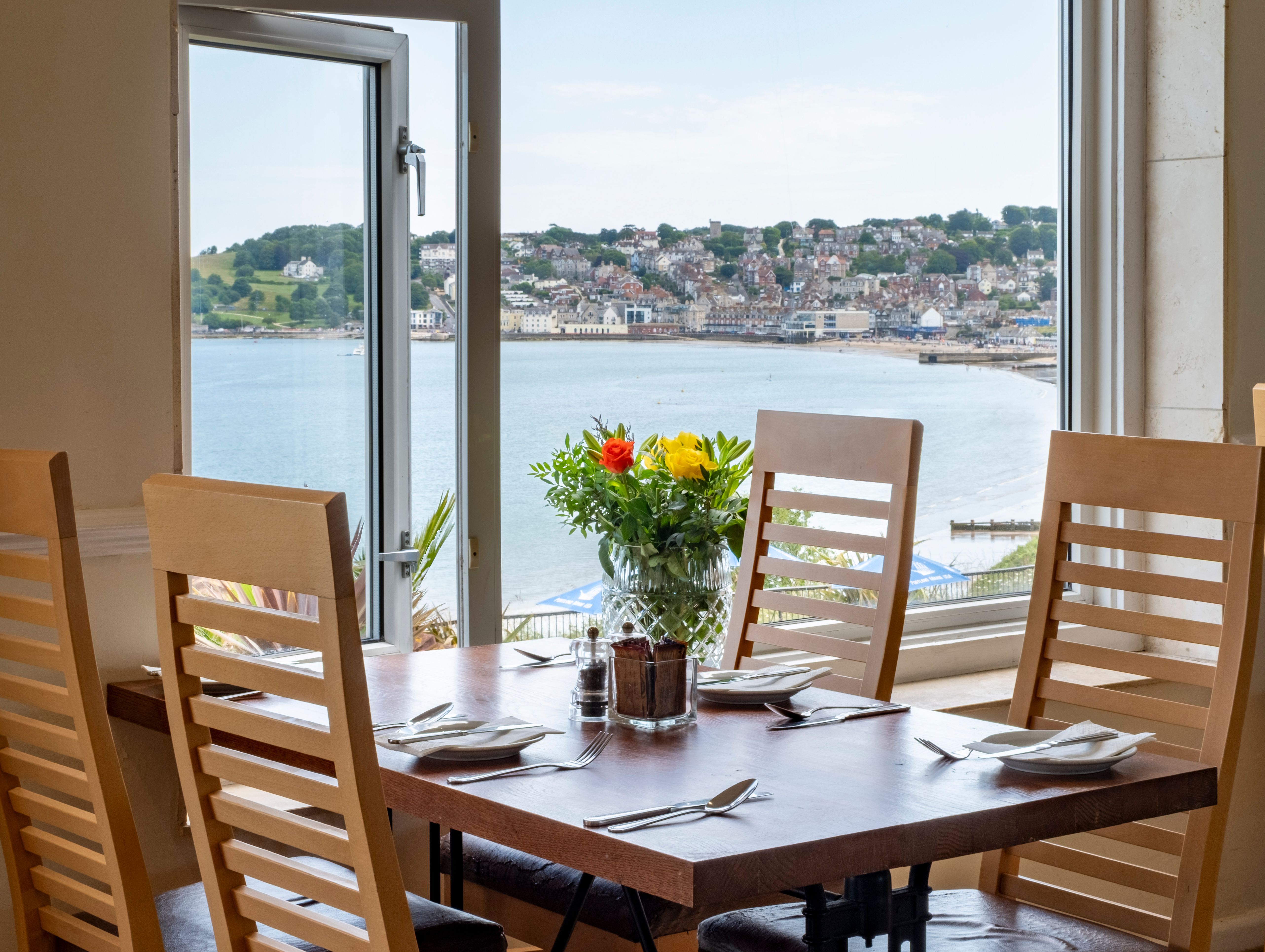 Dining table set by a large window with a scenic view of a coastal town and water, with flowers in a vase at the center of the table.