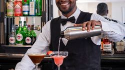 Bartender in a black vest and bow tie smiling while pouring a cocktail from a shaker into a glass at a bar, with various bottles and snacks visible on the counter.
