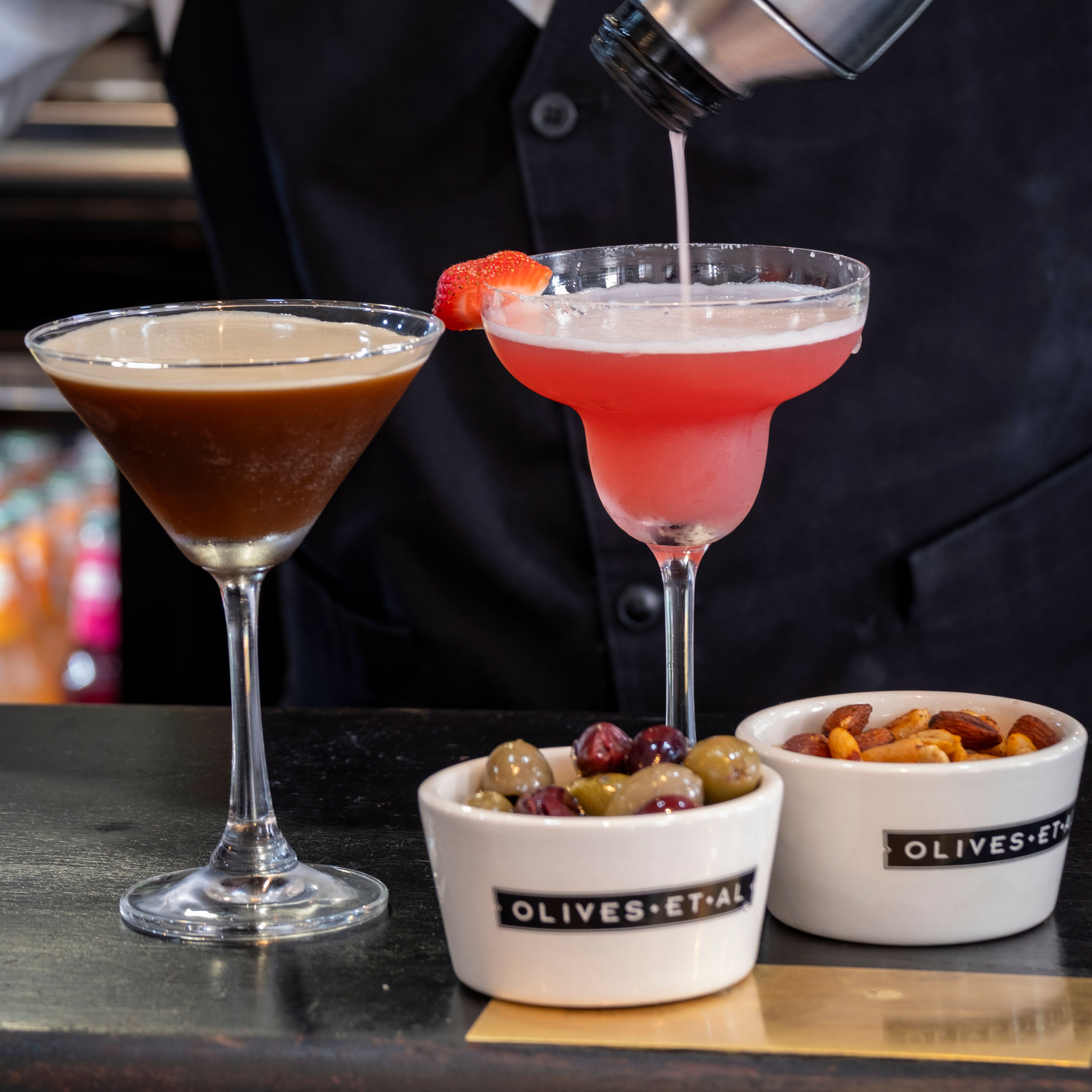Bartender pouring a pink cocktail from a shaker into a margarita glass, next to an espresso martini, a bowl of mixed olives, and a bowl of mixed nuts on a bar counter.