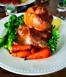 A plate of traditional roast beef dinner with Yorkshire pudding, roasted carrots, broccoli, sliced potatoes, and garnished with parsley.