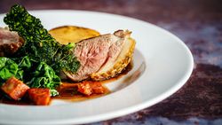 Gourmet plated dish featuring a slice of medium-rare meat, sautéed greens, crispy kale, roasted potato, and diced vegetables with sauce on a white plate.