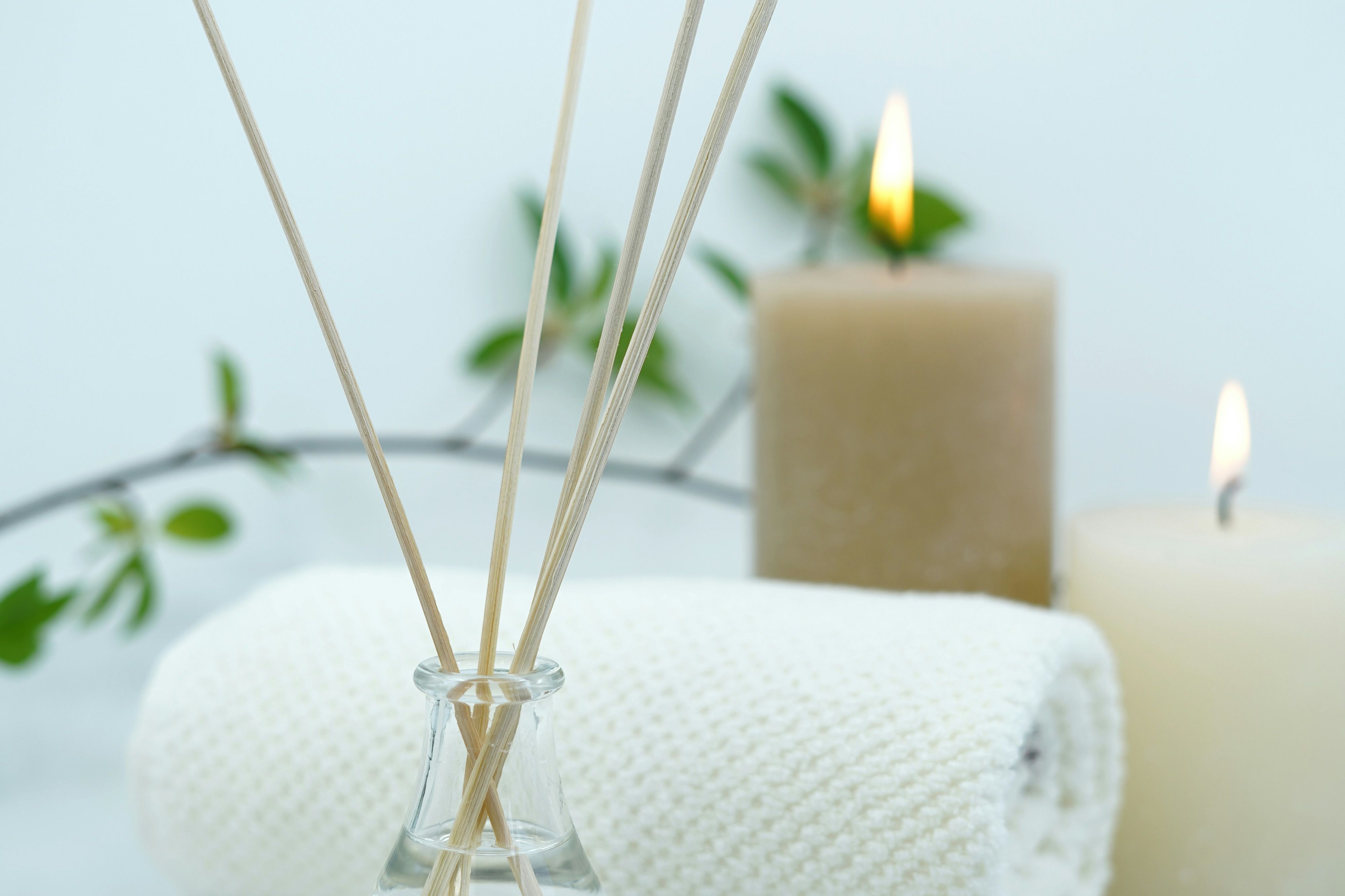 Aromatherapy sticks in a glass jar with two lit candles, a rolled white towel, and green leaves in the background, creating a spa-like ambiance.