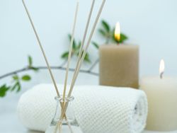 Aromatherapy sticks in a glass jar with two lit candles, a rolled white towel, and green leaves in the background, creating a spa-like ambiance.