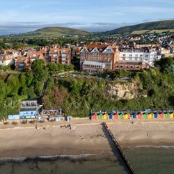 Aerial view of a seaside town with red-roofed buildings on a cliff, colorful beach huts along the sandy shore, and green hills in the background.