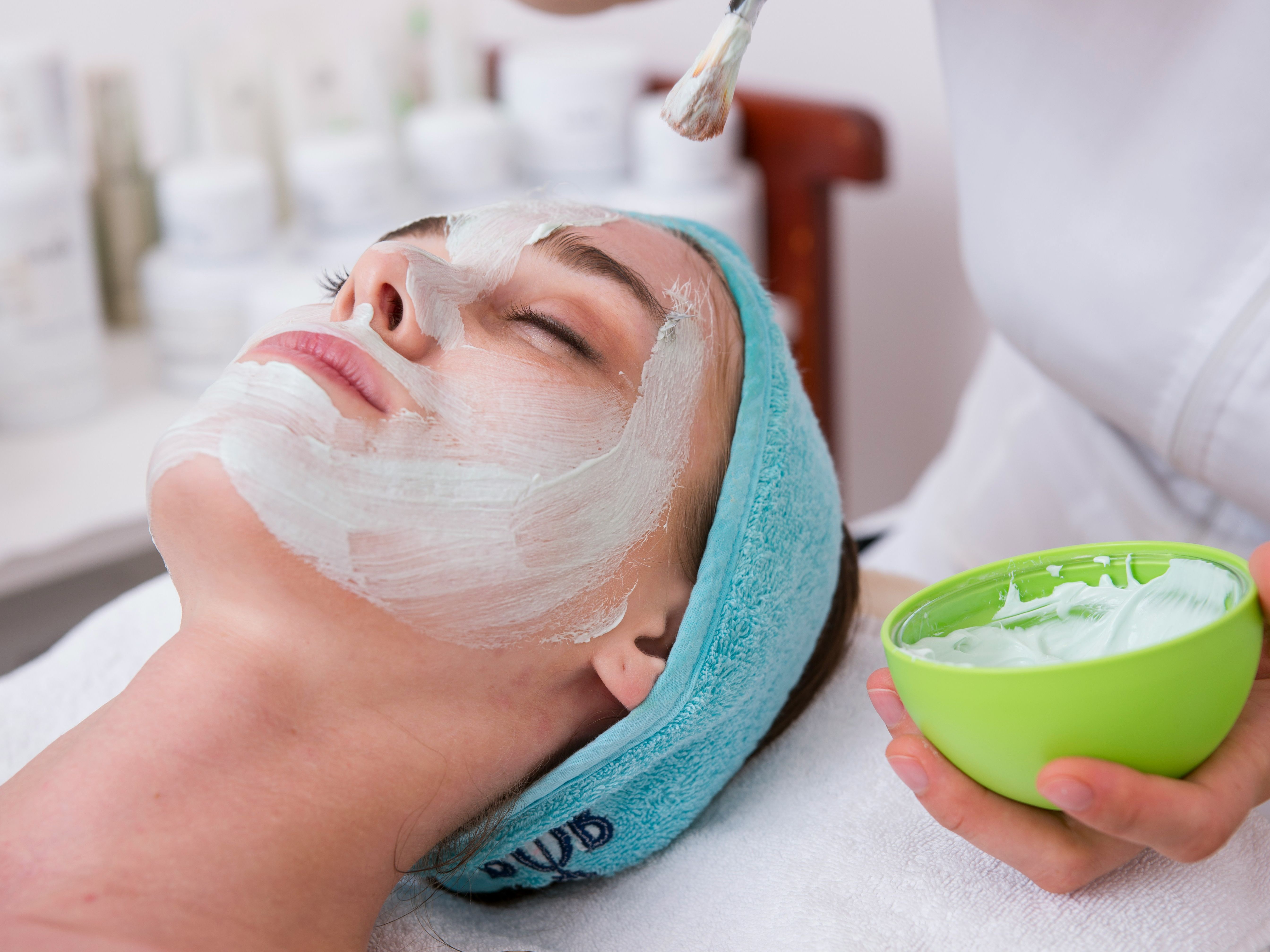 A woman receiving a facial treatment, lying down with a creamy facial mask being applied with a brush by a person holding a green bowl.