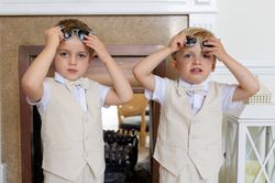 Two young boys in matching cream vests and bow ties adjusting sunglasses indoors.