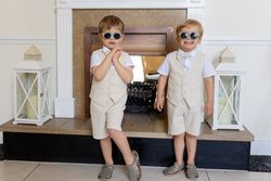 Two young boys dressed in beige shorts, vests, white shirts, and sunglasses standing in front of a fireplace.
