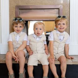 Three young boys dressed in matching beige vests and shorts sitting on a fireplace hearth, each wearing a bowtie and sunglasses.