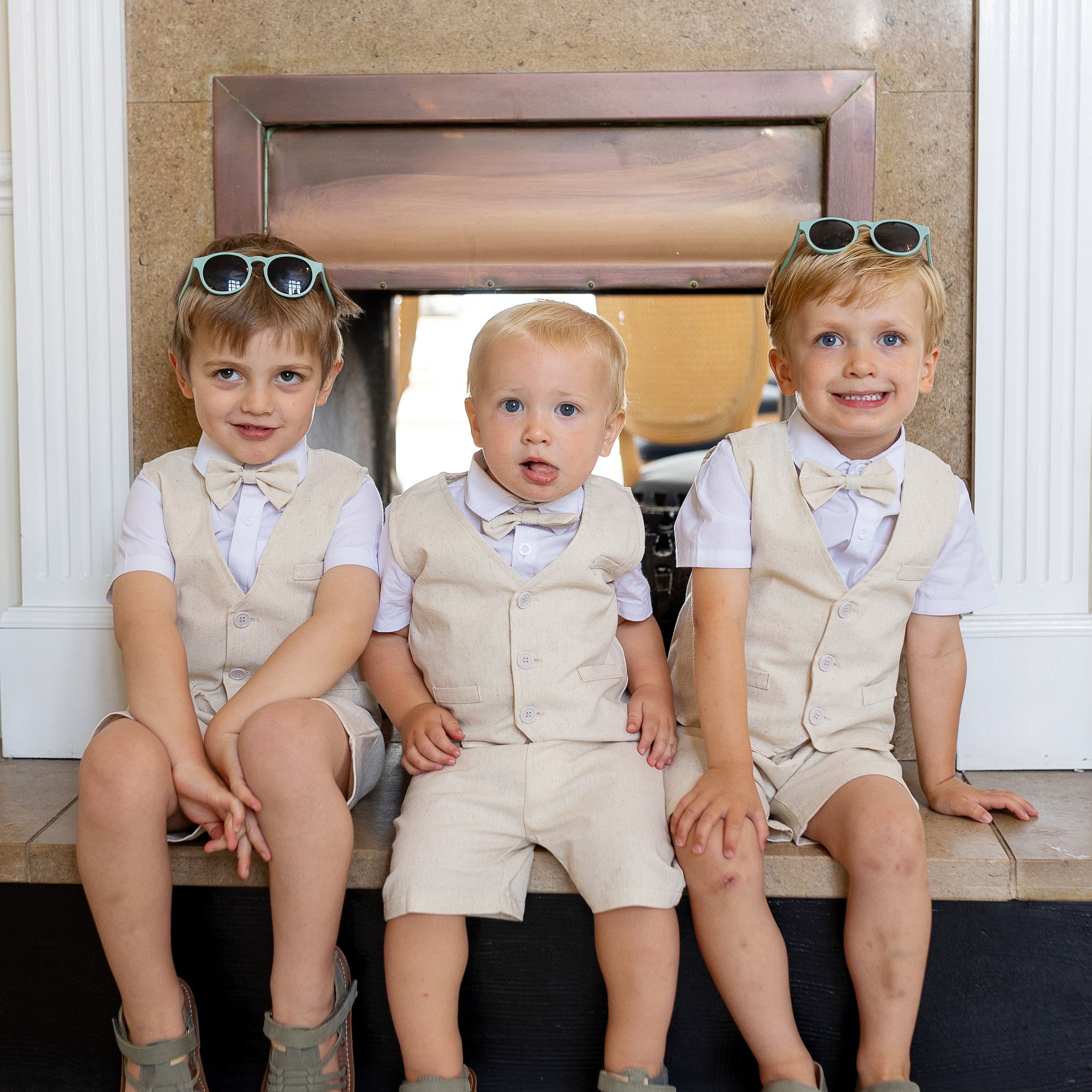 Three young boys dressed in matching beige vests and shorts sitting on a fireplace hearth, each wearing a bowtie and sunglasses.