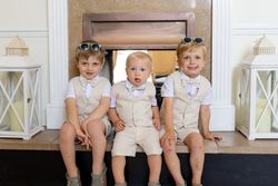 Three young boys dressed in matching beige vests and shorts sitting on a fireplace hearth, each wearing a bowtie and sunglasses.