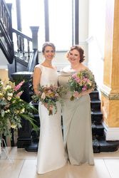 Two women in formal dresses holding bouquets, standing in front of a staircase.