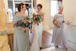 Three women dressed elegantly indoors, two holding bouquets and one holding a clutch bag and hat.