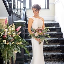 Bride in white gown holding bouquet standing on staircase with floral arrangement beside her
