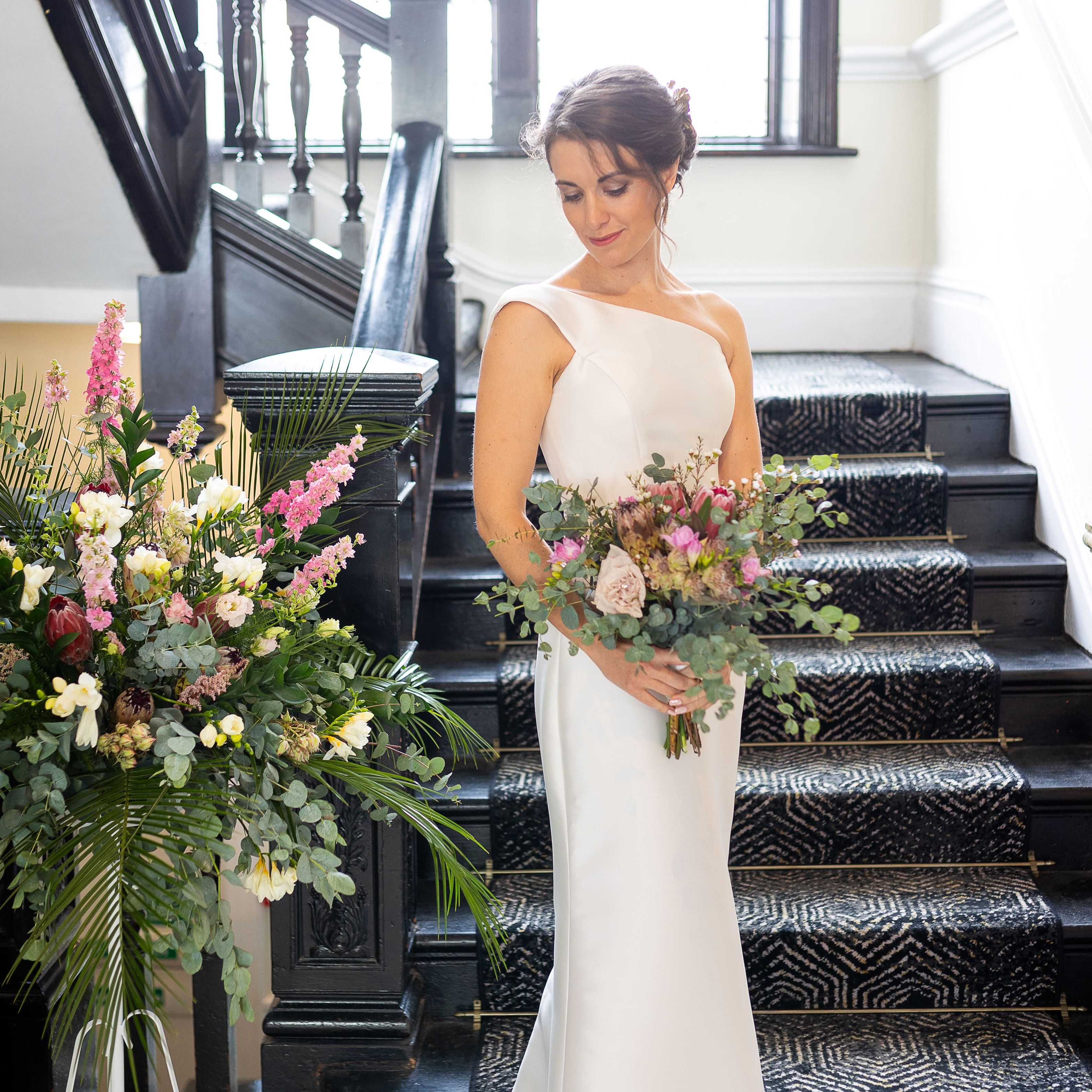 Bride in white gown holding bouquet standing on staircase with floral arrangement beside her
