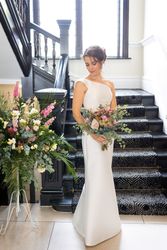 Bride in white gown holding bouquet standing on staircase with floral arrangement beside her
