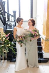 Two women in elegant dresses holding bouquets stand together by a staircase, smiling at each other.