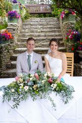 Smiling bride and groom sitting at a table with a large floral arrangement in an outdoor garden setting