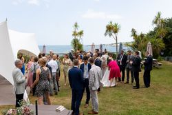 A group of people dressed formally, mingling outdoors at a wedding reception by the sea, with palm trees and white tent in the background.