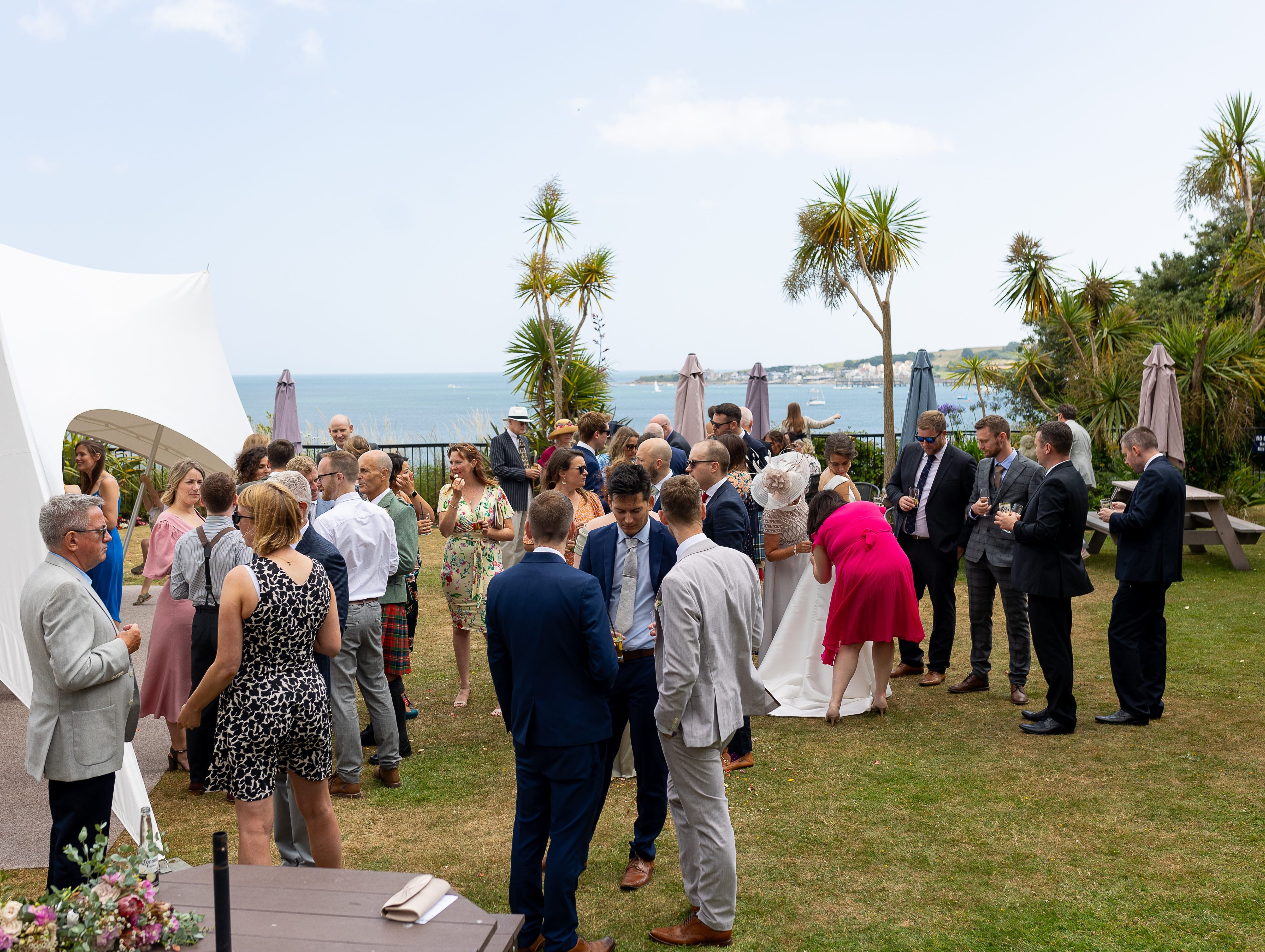 A group of people dressed formally, mingling outdoors at a wedding reception by the sea, with palm trees and white tent in the background.