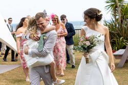 A bride and groom celebrating outdoors with wedding guests, the groom joyfully hugging a young girl.