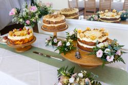 Three decorative cakes on wooden stands, surrounded by flowers and greenery on a white tablecloth.