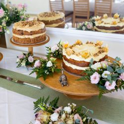 Three decorative cakes on wooden stands, surrounded by flowers and greenery on a white tablecloth.