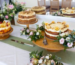 Three decorative cakes on wooden stands, surrounded by flowers and greenery on a white tablecloth.
