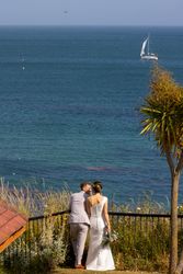 Bride and groom standing by a railing overlooking the sea with a sailboat in the distance.